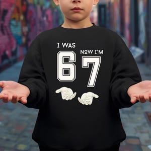 A person wearing a black sweatshirt with the text "I WAS 67 NOW I'M 67" printed on it.