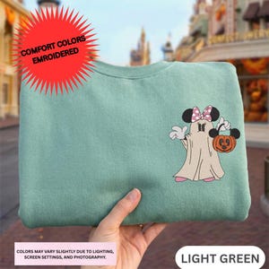 A hand holding a green sweatshirt with a Minnie Mouse and a ghost character printed on it.
