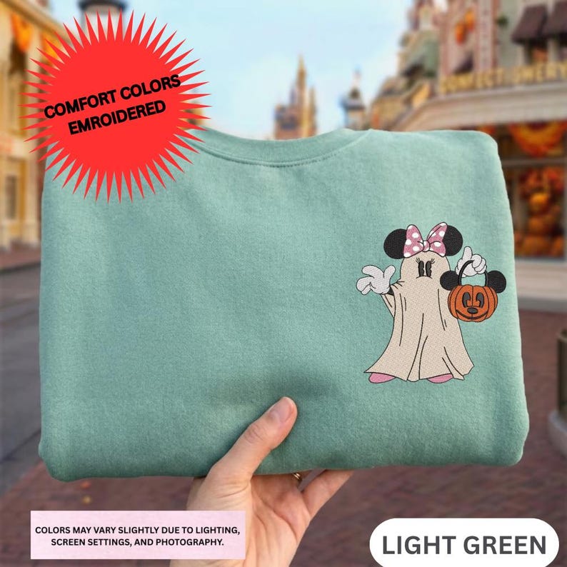 A hand holding a green sweatshirt with a Minnie Mouse and a ghost character printed on it.