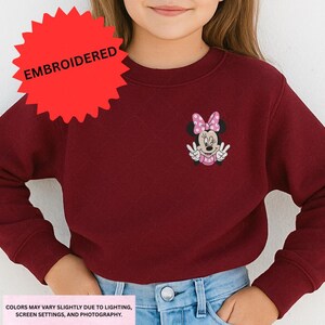 A young girl wearing a burgundy sweater with a Minnie Mouse embroidered on it.