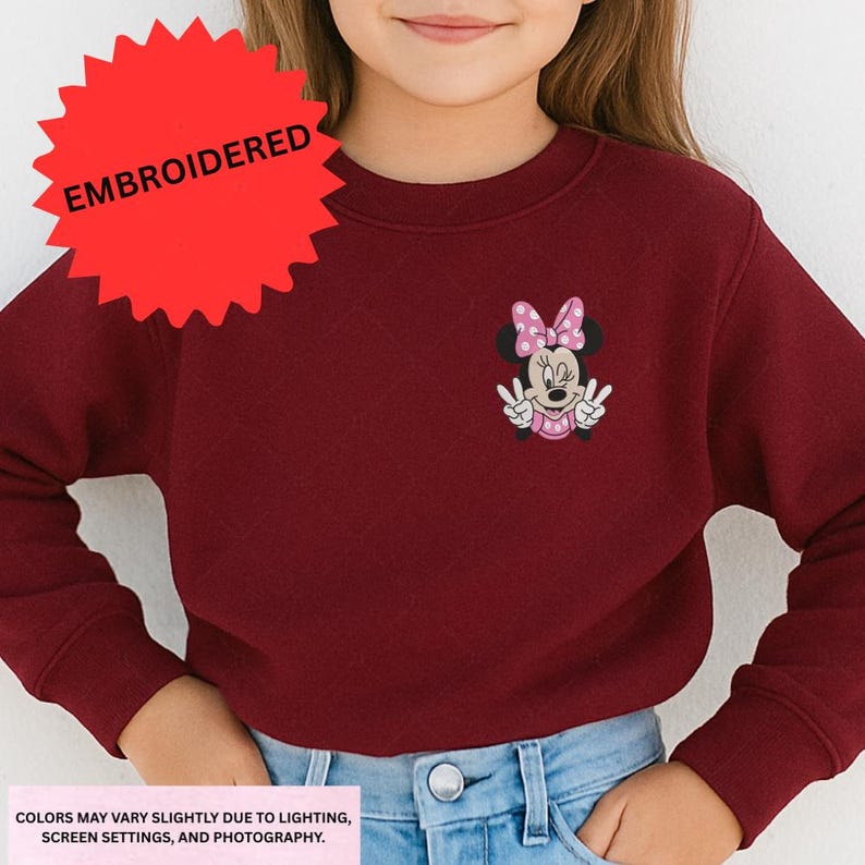 A young girl wearing a burgundy sweater with a Minnie Mouse embroidered on it.