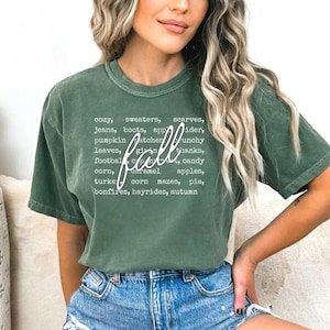 May include: A green t-shirt with the word "fall" in cursive script. The shirt lists autumn-themed words like "cozy," "sweaters," "pumpkin," and "bonfires" in a white, sans-serif font.