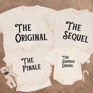 May include: Four cream-colored t-shirts and a onesie with black text. The shirts read "The Original", "The Sequel", "The Finale", and the onesie says "The Surprise Ending". A small stuffed bear and a string of beads are also visible.