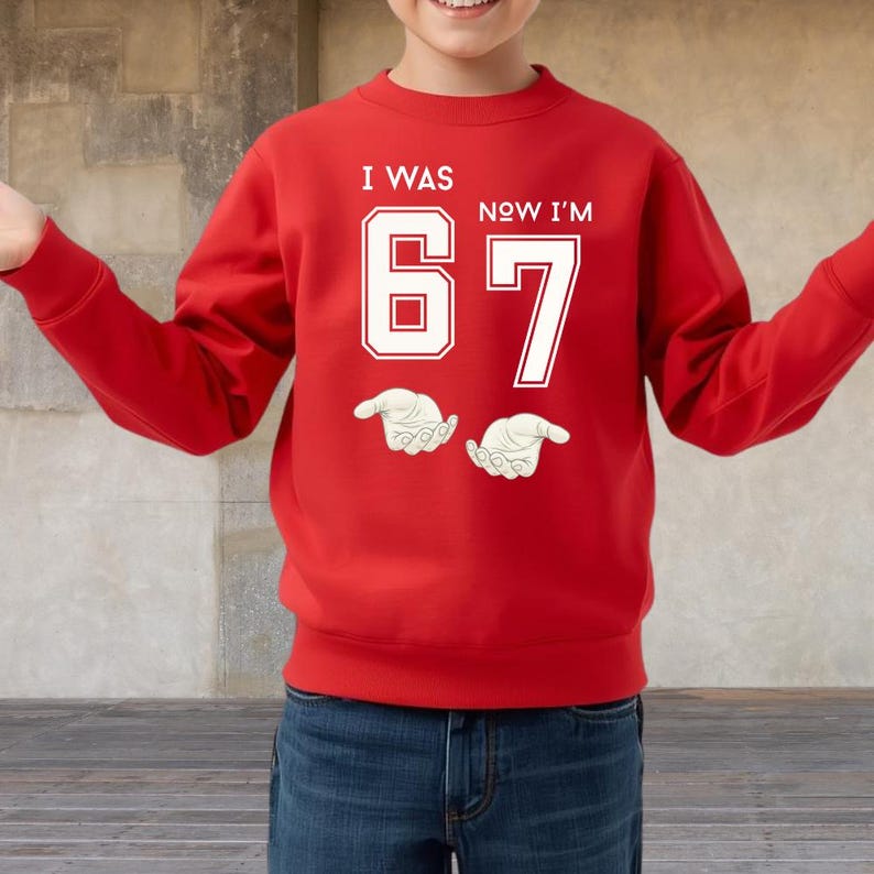 A person wearing a red sweatshirt with the text "I WAS 67 NOW I'M" printed on it.