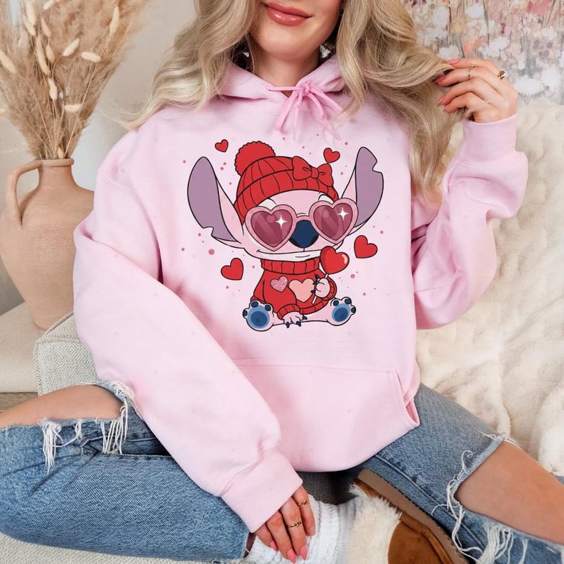 A woman wearing a pink hoodie with a cartoon character and heart-shaped glasses is sitting on a couch.