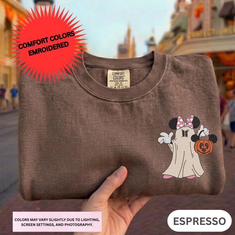 A brown t-shirt with a Minnie Mouse and a ghost character printed on it, along with a red starburst logo and text indicating that the colors may be slightly faded and that the shirt is embroidered.