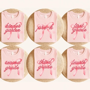 Puede incluir: Seis camisetas de color rosa claro con diferentes textos de nivel de grado en fuente cursiva y un gráfico de cinta rosa. Los textos incluyen "kindergarten", "pre-k", "first grade", "second grade", "third grade" y "fourth grade".