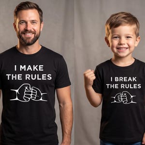 May include: Two people wearing matching black t-shirts. The adult's shirt says "I MAKE THE RULES" with a graphic of two fists bumping. The child's shirt says "I BREAK THE RULES" with a similar graphic.