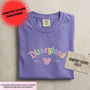 a purple t-shirt with the Disneyland logo and text, along with a tag that says "Comfort Colors Violet" and a red starburst that says "COMFORT COLORS EMBROIDED".