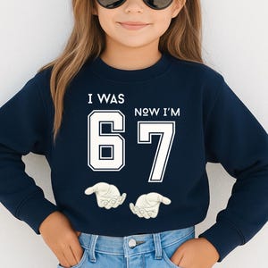 A young girl wearing a navy blue sweatshirt with the text "I WAS 67 NOW I'M 67" printed on it.