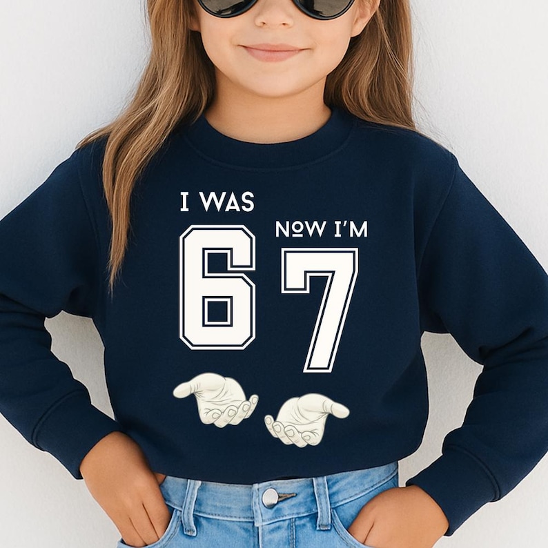 A young girl wearing a navy blue sweatshirt with the text "I WAS 67 NOW I'M 67" printed on it.