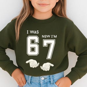A young girl wearing a green sweatshirt with the text "I WAS 67 NOW I'M" printed on it.