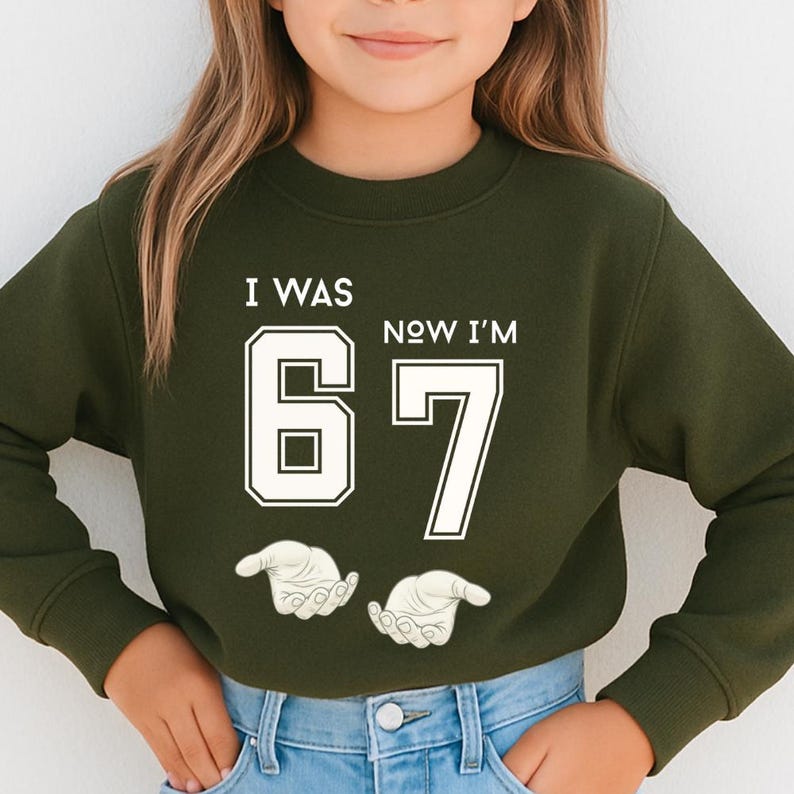 A young girl wearing a green sweatshirt with the text "I WAS 67 NOW I'M" printed on it.
