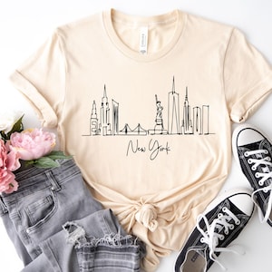 May include: A light beige short-sleeved t-shirt featuring a black continuous line drawing of the New York City skyline, including the Statue of Liberty, with the text New York written in a cursive script font centered below the graphic.