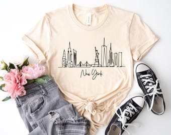 May include: A light beige short-sleeved t-shirt featuring a black continuous line drawing of the New York City skyline, including the Statue of Liberty, with the text New York written in a cursive script font centered below the graphic.