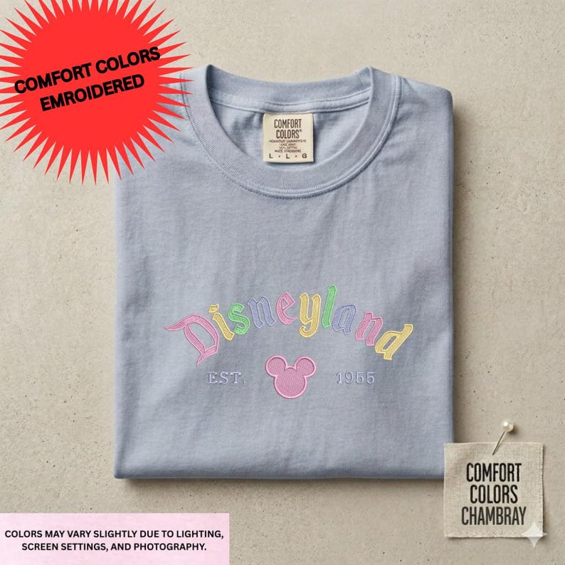 a gray t-shirt with the text "Disneyland" and "EST. 1955" embroidered on it, along with a Mickey Mouse logo. The t-shirt is displayed on a flat surface, and there is a red starburst and a label with the text "COMFORT COLORS" and "EMROIDERED" on it.
