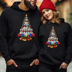 A man and a woman are standing next to each other, both wearing black hooded sweatshirts with a Christmas tree design.