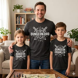 May include: Three people wearing matching dark gray t-shirts with white graphic designs. The adult's shirt says "DUNGEON DAD" with crossed swords and a die. The children's shirts say "DUNGEON CRAWLER" with similar graphics. They are standing in front of a table with game pieces.