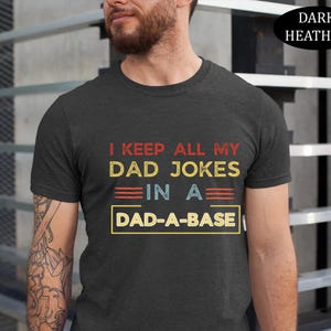 May include: Dark heather gray t-shirt with the text "I KEEP ALL MY DAD JOKES IN A DAD-A-BASE" in red, yellow, and blue. The shirt is a crew neck style.