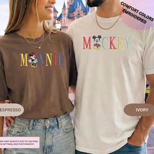 A woman wearing a brown t-shirt with the word "Minnie" and a man wearing a white t-shirt with the word " Mickey" printed on them, standing together in front of a castle.