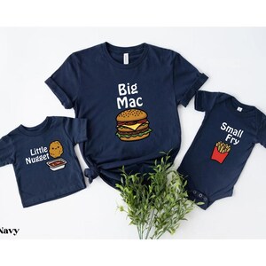 May include: Navy blue t-shirts and a onesie with food-themed graphics. The adult shirt says "Big Mac" with a burger illustration. The child's shirt says "Little Nugget" and the onesie says "Small Fry".