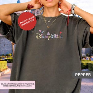 A person wearing a gray t-shirt with the Disney World logo, posing with their hands on their ears.