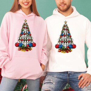 A woman wearing a pink hoodie with a Christmas tree design and a man wearing a white hoodie with a Christmas tree design.