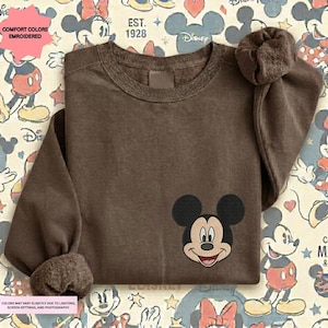 A brown t-shirt with a Mickey Mouse embroidered on the front, placed on a background with various Disney characters and logos.
