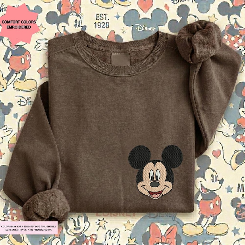 A brown t-shirt with a Mickey Mouse embroidered on the front, placed on a background with various Disney characters and logos.