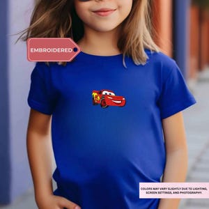 A young girl wearing a blue t-shirt with a red and yellow cartoon car design on it.