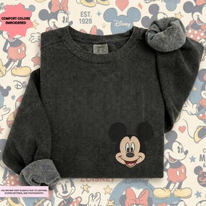 a black sweater with a Mickey Mouse embroidered on the front, placed on a background with Disney-themed graphics.