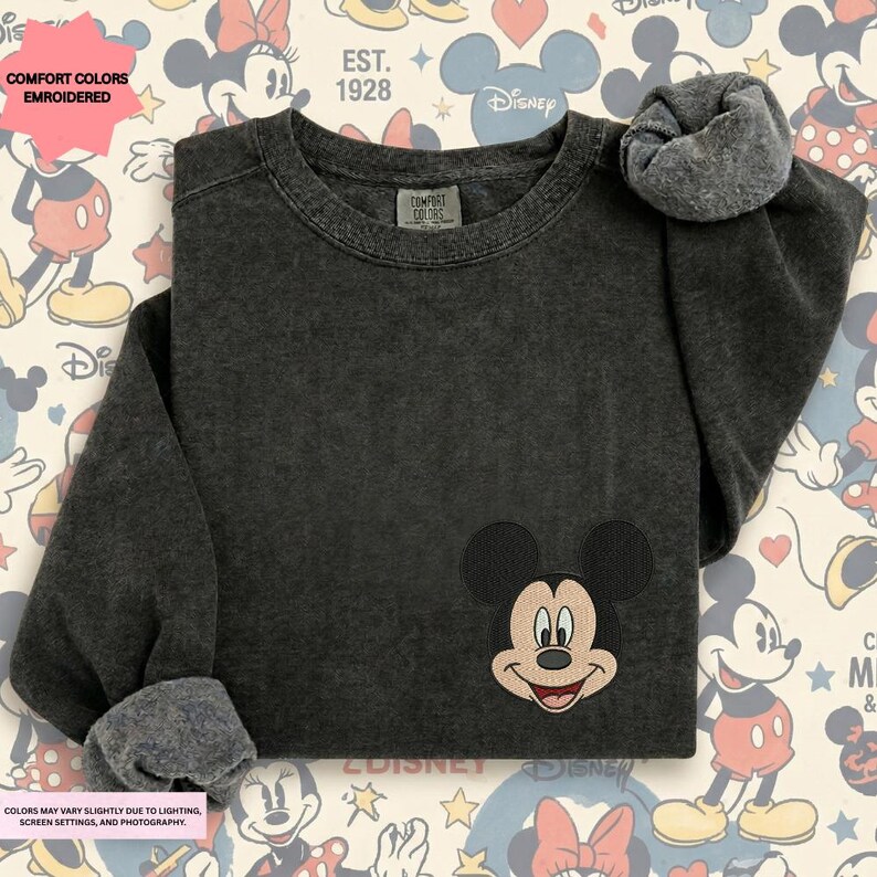 a black sweater with a Mickey Mouse embroidered on the front, placed on a background with Disney-themed graphics.