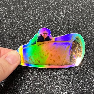 May include: A holographic sticker of a man dressed in a black shirt, sitting inside a large burrito. The sticker has a rainbow iridescent finish.