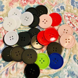 May include: A collection of round, four-hole buttons in various solid colors including black, white, red, light pink, lime green, and royal blue. The buttons have a smooth, glossy finish and are arranged in a scattered, overlapping cluster.
