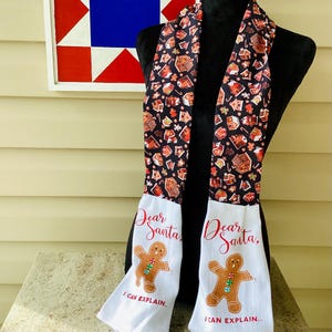 May include: A hanging kitchen towel featuring a black fabric top patterned with gingerbread houses and candy canes. The white bottom section displays a gingerbread man graphic with the red text "Dear Santa, I CAN EXPLAIN.." printed above and below the figure.