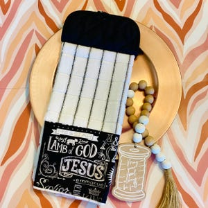 May include: A hanging kitchen towel featuring a white and gray checkered pattern with a black quilted top and a black bottom panel printed with white text including Messiah, Lamb of God, Jesus, Emmanuel, and Savior, alongside a wooden spool ornament.