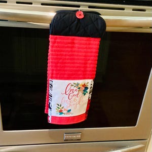 May include: A red and black oven mitt and towel set. The black mitt has a quilted pattern and a red button. The red towel has the words "Love God Love & People" with floral accents.