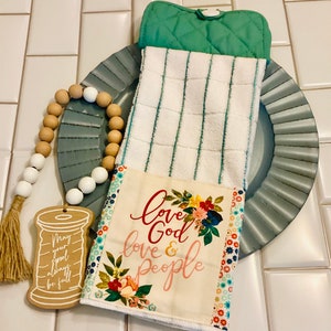 May include: A white kitchen towel with green stripes and a floral print panel that says "Love God & Love People". The towel is folded and placed on a blue plate with a wooden bead garland.