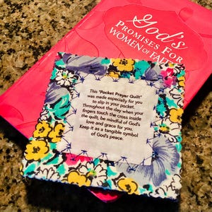 May include: A small, square Pocket Prayer Quilt with floral patterns in yellow, blue, and pink, with text. The quilt is placed on top of a pink book titled "God's Promises for Women of Faith."