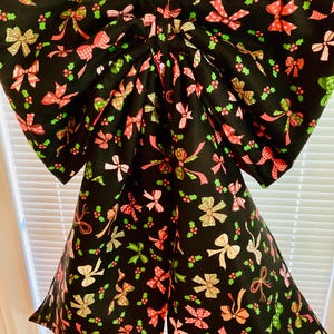 May include: A large fabric bow featuring a black background with a repeating pattern of small colorful bows in shades of red, pink, green, and white, interspersed with tiny green holly leaves and red berries, suspended from a silver metal ring.