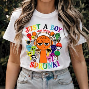 May include: White t-shirt with a colorful graphic design featuring a cartoon character with headphones and the text "Just a boy who loves Sprunki".