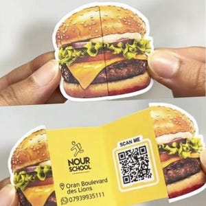 Può includere: Un adesivo a forma di hamburger, con panino al sesamo, lattuga, pomodoro, formaggio e un hamburger. L'adesivo si apre per rivelare un interno giallo con la scritta "NOUR SCHOOL", un codice QR e informazioni di contatto.