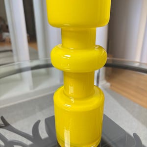 Peut inclure: Un vase en verre jaune vif avec un design unique empil&eacute;. Le vase pr&eacute;sente une base cylindrique, une section m&eacute;diane arrondie et un dessus large et rectangulaire. La finition brillante refl&egrave;te la lumi&egrave;re, soulignant la couleur vibrante et la texture lisse. (Environ 20 cm de haut)