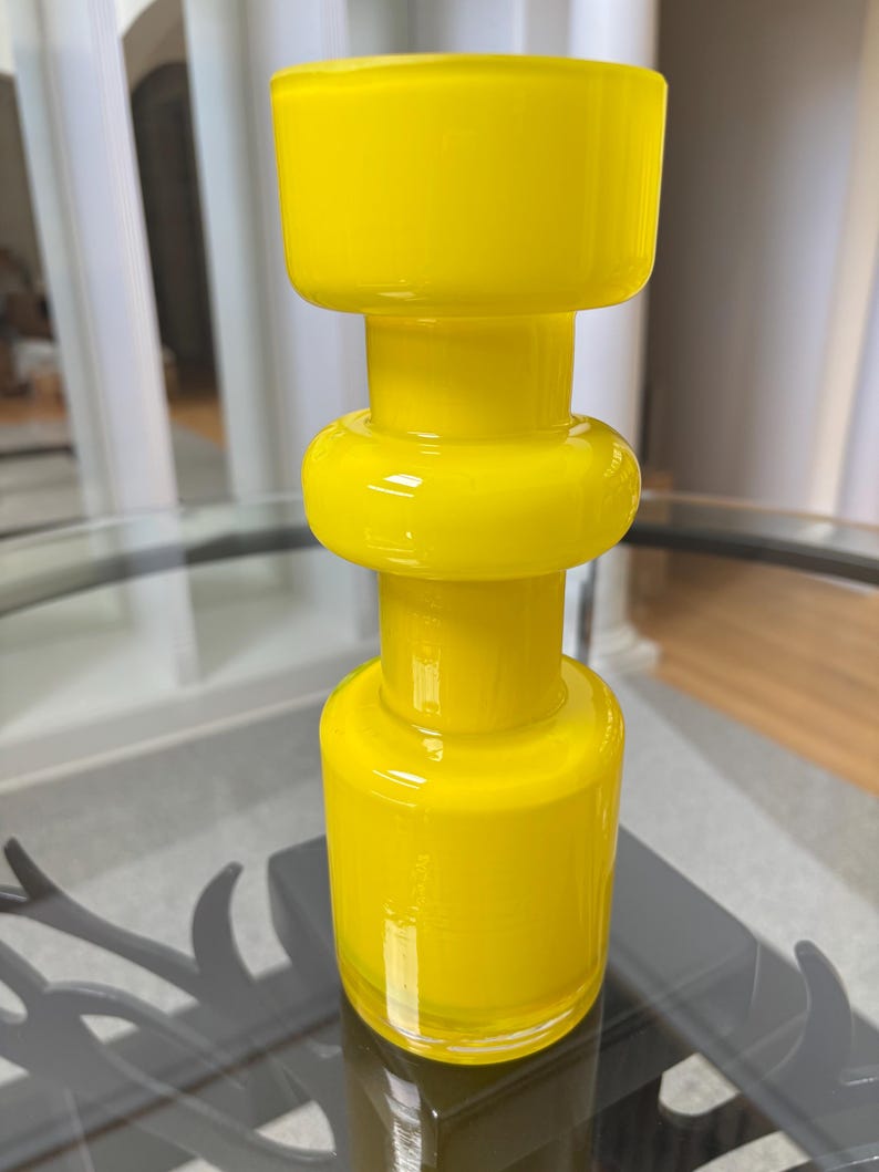 Peut inclure: Un vase en verre jaune vif avec un design unique empil&eacute;. Le vase pr&eacute;sente une base cylindrique, une section m&eacute;diane arrondie et un dessus large et rectangulaire. La finition brillante refl&egrave;te la lumi&egrave;re, soulignant la couleur vibrante et la texture lisse. (Environ 20 cm de haut)