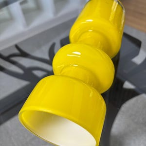 Peut inclure: Une lampe en verre jaune vif au design unique et empil&eacute;. La lampe pr&eacute;sente un dessus et un dessous cylindriques, reli&eacute;s par une section centrale arrondie. L'int&eacute;rieur de la lampe est blanc, cr&eacute;ant un contraste avec l'ext&eacute;rieur jaune vif.