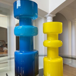 Peut inclure: Deux vases en verre, un bleu et un jaune, sont expos&eacute;s sur une table en verre. Le vase bleu pr&eacute;sente un d&eacute;grad&eacute; de bleu fonc&eacute; &agrave; bleu clair, avec une bande argent&eacute;e &agrave; la base. Le vase jaune est de couleur unie.