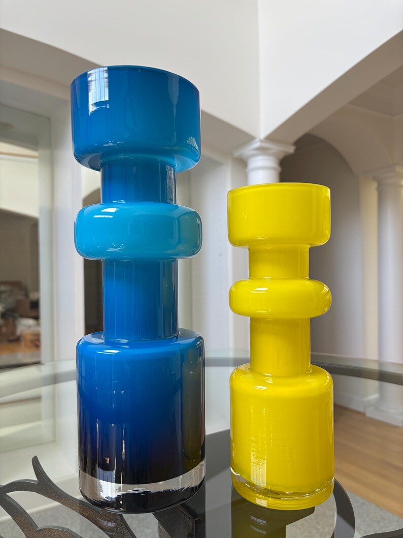 Peut inclure: Deux vases en verre, un bleu et un jaune, sont expos&eacute;s sur une table en verre. Le vase bleu pr&eacute;sente un d&eacute;grad&eacute; de bleu fonc&eacute; &agrave; bleu clair, avec une bande argent&eacute;e &agrave; la base. Le vase jaune est de couleur unie.