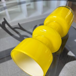 Peut inclure: Un vase en verre jaune vif avec un design empil&eacute; unique. Le vase pr&eacute;sente une base cylindrique, deux sections m&eacute;dianes arrondies et un sommet &eacute;vas&eacute;. L'int&eacute;rieur de la section sup&eacute;rieure est blanc, cr&eacute;ant un contraste. Le vase est sur une table en verre.