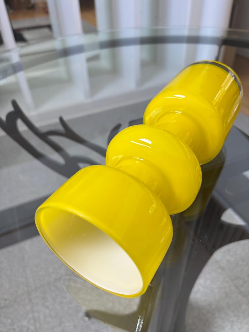 Peut inclure: Un vase en verre jaune vif avec un design empil&eacute; unique. Le vase pr&eacute;sente une base cylindrique, deux sections m&eacute;dianes arrondies et un sommet &eacute;vas&eacute;. L'int&eacute;rieur de la section sup&eacute;rieure est blanc, cr&eacute;ant un contraste. Le vase est sur une table en verre.