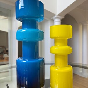 Peut inclure: Deux vases d&eacute;coratifs en verre, l'un bleu avec une base argent&eacute;e, l'autre jaune. Les deux ont une base cylindrique, une section centrale arrondie et un large dessus plat. Ils sont pos&eacute;s sur une table en verre.